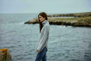 bellissimo donna nel un' grigio maglione sta su un' roccioso riva natura stile di vita foto