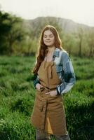 un' contadino donna nel sua opera Abiti, plaid camicia e grembiule, sta nel il campo su il verde erba e sorrisi nel il ambientazione sole dopo un' difficile giorno opera foto