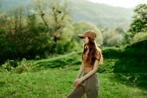 un' bellissimo donna nel abbigliamento sportivo e un' berretto passeggiate contro un' verde naturale paesaggio e sorrisi nel il luce del sole foto