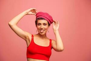 giovane atletico donna con un' corto taglio di capelli e viola capelli nel un' rosso superiore e un' rosa cappello con un atletico figura sorrisi e smorfie guardare a il telecamera su un' rosa sfondo foto