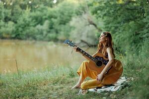 un' hippie donna giocando sua chitarra sorrisi e canta canzoni nel natura seduta su un' plaid nel il sera nel il tramonto luce del sole. un' stile di vita nel armonia con il corpo e natura foto