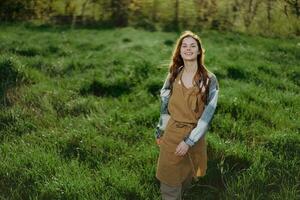 un' donna giardiniere nel un grembiule sta nel un' campo di verde erba all'aperto, sorridente su un' estate pomeriggio in un' soleggiato tramonto dopo un' giorno opera foto