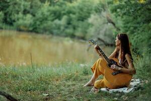 un' hippie donna giocando sua chitarra sorrisi e canta canzoni nel natura seduta su un' plaid nel il sera nel il tramonto luce del sole. un' stile di vita nel armonia con il corpo e natura foto