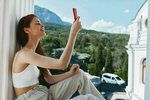 bellissimo donna seduta su il balcone con Telefono bellissimo montagna Visualizza estate rilassamento concetto foto