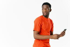 un africano uomo nel un arancia maglietta sembra indietro e detiene un' mobile Telefono nel il suo mano foto