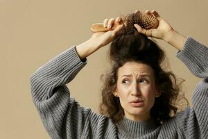 problematico indisciplinato danneggiato capelli concetto. sofferenza Riccio bellissimo donna nel grigio casuale maglione con spazzola per capelli pettine tira ingarbugliato capelli su in posa isolato su al di sopra di beige pastello sfondo. copia spazio foto