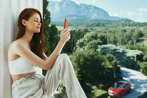 attraente giovane donna seduta su il balcone con Telefono bellissimo montagna Visualizza estate inalterato foto