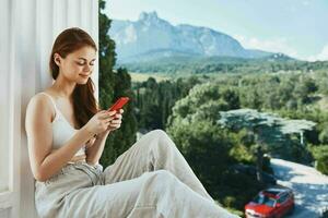 ritratto di bellissima donna seduta su il balcone con Telefono bellissimo montagna Visualizza estate rilassamento concetto foto