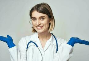 un' infermiera nel un' medico toga e blu guanti gesti con un' stetoscopio in giro sua collo foto