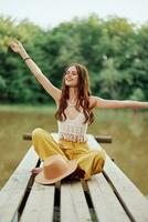 un' hippie donna si siede su un' ponte di un' lago con sua mani su nel il aria mentre in viaggio nel natura e sorridente nel eco-abbigliamento. rilassato stile di vita foto