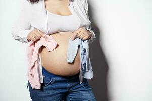 avvicinamento incinta donna Tenere blu e rosa neonato body vicino sua gonfiarsi. aspettandosi per un' bambino, ragazzo o ragazza, Gemelli foto