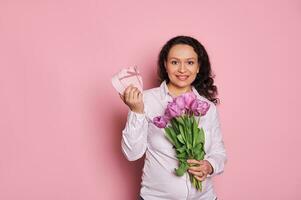 bellissimo donna mostrando a telecamera un' carino regalo scatola, in posa con mazzo di tulipani per festivo occasione al di sopra di rosa sfondo foto