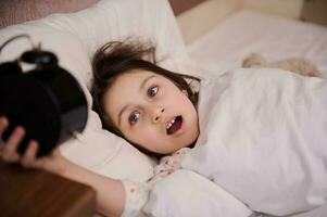 adorabile bambino ragazza dire bugie nel letto su cuscino, veglia su e commutazione via allarme orologio nel il presto mattina foto