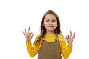 malizioso sorridente poco bambino ragazza mostrando ok cartello, esprimendo felicità e positivo emozioni al di sopra di bianca sfondo foto