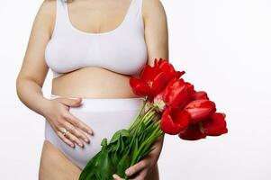 avvicinamento corpo positivo incinta donna nel biancheria intima, mettendo mano su gonfiarsi, in piedi con mazzo di tulipani su bianca fondale foto