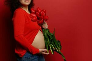 avvicinamento di incinta pancia con allungare votazione di un' donna, in attesa madre in posa con rosso tulipani su isolato sfondo foto