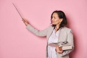 fiducioso mezzo anziano donna, scuola insegnante professoressa, punti pointer a copia spazio su rosa parete mentre spiegando lezione foto