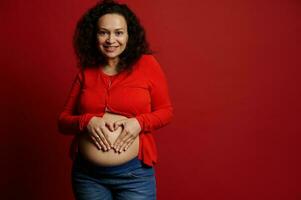 affascinante incinta donna mostrando un' cuore fatto a partire dal sua dita su sua gonfiarsi, sorridente guardare a telecamera, isolato su rosso foto