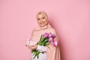 sorridente medio orientale musulmano donna nel hijab, con tulipani e regalo scatola su rosa sfondo . internazionale Da donna giorno foto