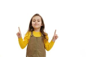 caucasico poco bambino ragazza punti dito su, trova soluzione, sorrisi guardare a telecamera, bianca sfondo. indietro per scuola foto