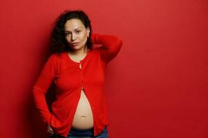 attraente bellissimo incinta donna, in attesa madre, guardare a telecamera, vestito nel rosso camicia, mostrando sua spoglio pancia foto
