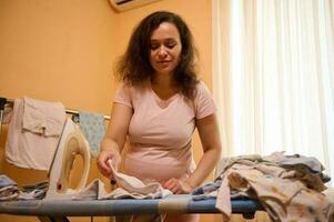 in attesa madre, incinta donna Stirare e pieghevole neonato Abiti, godendo futuro maternità e spensierato gravidanza foto