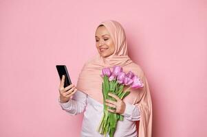 musulmano donna nel rosa hijab, utilizzando smartphone, scorrimento notizia alimentazione, controllo sociale media contenuto, analisi nuovo mobile App foto