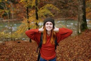 gioioso donna nel un' cappello maglione con un' zaino su sua indietro gesti con sua mani nel un' parco nel natura foto