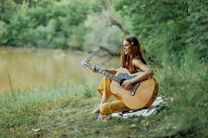 hippie donna giocando chitarra sorridente e cantando canzoni nel natura seduta su un' plaid di il lago nel il sera nel il raggi di il ambientazione sole. un' stile di vita nel armonia con il corpo e natura foto