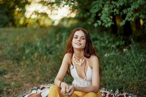 ragazza vestito come un' hippie eco rilassante nel il parco, seduta su un' coperta nel il tramonto, rilassato stile di vita foto