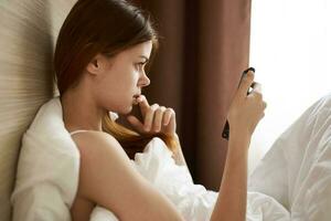 un' donna sotto il coperture bugie nel letto e sembra a il mobile Telefono schermo lato Visualizza foto