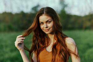 un' giovane donna sorrisi e sembra in il telecamera con sua lungo, ondulato, brillante rosso capelli nel un' parco con verde erba nel il estate tramonto foto