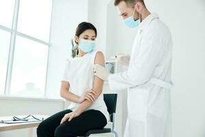 un' medico nel un' medico toga e un' maschera inietta un' femmina paziente nel il spalla covid vaccinazione foto