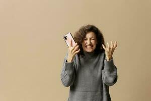 il male irritata Riccio bellissimo femmina nel grigio casuale maglione spremere Telefono con furia in posa isolato su al di sopra di beige pastello sfondo. sociale media, Rete, distanza comunicazione concetto. copia spazio foto