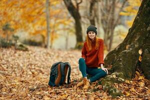donna nel i pantaloni e stivali con un' zaino si siede vicino un' albero nel il autunno foresta caduto le foglie foto