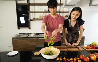 una giovane coppia asiatica sta mangiando insieme e sorridendo felicemente mentre cucina la loro insalata in cucina. foto