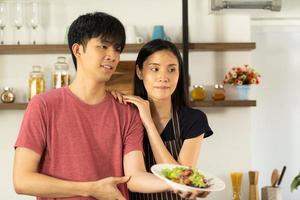 una giovane coppia asiatica sta mangiando insieme e sorridendo felicemente mentre cucina la loro insalata in cucina. foto