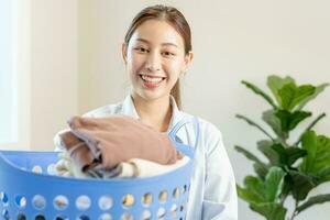 casalinga, attraente asiatico giovane donna, ragazza Tenere molti pieghevole Abiti nel il cestino dopo lavaggio pulito Abiti e essiccazione, domestico Lavorando nel camera a casa, domestica lavoretto lavanderia ordinato. foto