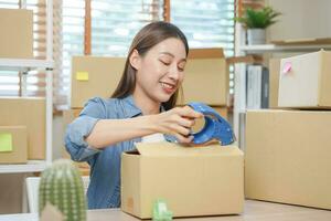 piccolo attività commerciale imprenditore me, asiatico giovane donna, ragazza proprietario Imballaggio Prodotto, controllo pacco per consegna per cliente, utilizzando scotch nastro per foca il scatola, Lavorando a casa. mercante in linea, e commercio foto