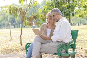 coppia di anziani caucasici si siede e si rilassa su una panchina nel parco e utilizza un computer portatile. concetto di vivere la vita felicemente in vacanza