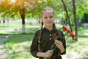 un' ragazza nel un' speciale uniforme con un' st. Giorgio nastro detiene tre garofani nel sua mani. vacanza vittoria giorno Maggio 9°. foto