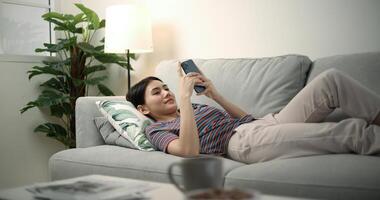 giovane asiatico femmina sorridente e fare surf il Internet su un' mobile Telefono mentre dire bugie su il divano foto