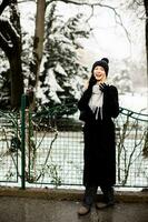 sorridente giovane donna nel accogliente capi di abbigliamento utilizzando mobile Telefono e Tenere caffè tazza su inverno giorno foto
