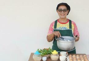 contento asiatico anziano donna è cucinando, indossare capocuoco berretto e grembiule, detiene pentola e mestolo. concetto, cucinando per famiglia. cucinando per vendere. tailandese cucina stile di vita. anziano attività. foto