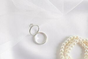 Due bianca oro anelli con un' diamante su il parte di il della sposa nozze velo e raso bianca tessile con perla perline. nozze composizione. foto