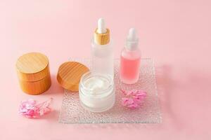 un' alla moda cosmetico Prodotto nel bicchiere Opaco bianca bottiglie e crema nel un' vaso su un' bicchiere sbalzato vassoio e un' rosa sfondo. naturale cosmetici. foto