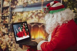 contento Santa Claus fa video chiamata bambino piccolo parlando per bambino, saluto allegro Natale, contento nuovo anno nel virtuale video in linea Chiacchierare su computer portatile, seduta nel davanti di il il camino. foto