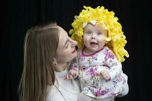 mamma detiene nel sua braccia un' poco contento figlia nel un' giallo parrucca. divertente bambini. foto