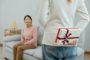 asiatico adolescente ragazze siamo nascondiglio regalo scatole per loro bellissimo mamme dietro a loro schiene mentre mamma è seduta su il divano a casa. festeggiare speciale occasione insieme a casa, contento compleanno per mamma foto