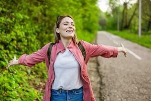 contento donna è pronto per escursioni a piedi nel foresta. lei è in piedi accanto il strada con braccia proteso. foto